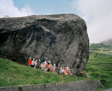 Scarecrows dressed in clothing and hats stand in a row beneath a huge boulder on a hillside