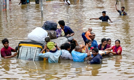 A group of people wading through brown, waist-high flood water clinging to a vehicle with their belongings on its roof