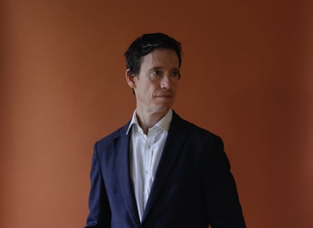 Rory Stewart wearing a navy suit and white shirt against a brown background