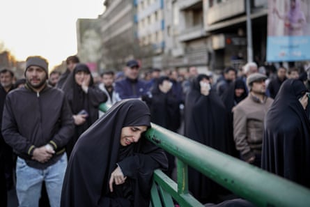A woman in black hijab leans against a railing in grief. Other people are out of focus behind