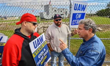 Standing in front of a chain-link fenced topped with barbed wire, beyond which is a massive freestanding 3D Jeep sign/struture, stand three men, all white-presenting. On the left are two men in sunglasses and baseball caps holding blue-and-white UAW picket signs, and on the left is a middle-aged man with gray curly hair and a blue denim shirt raising his hand and pointing as one of the men, although he appears to be making a point, not threatening him.