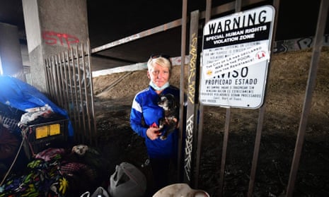 Dawn Wilson, with her dog, Lily, stands at a homeless encampment beneath a freeway overpass near SoFi Stadium on Wednesday. She stands next to a sign warning against camping that has been covered with a sticker saying 'the city of LA has declared war on the poor'