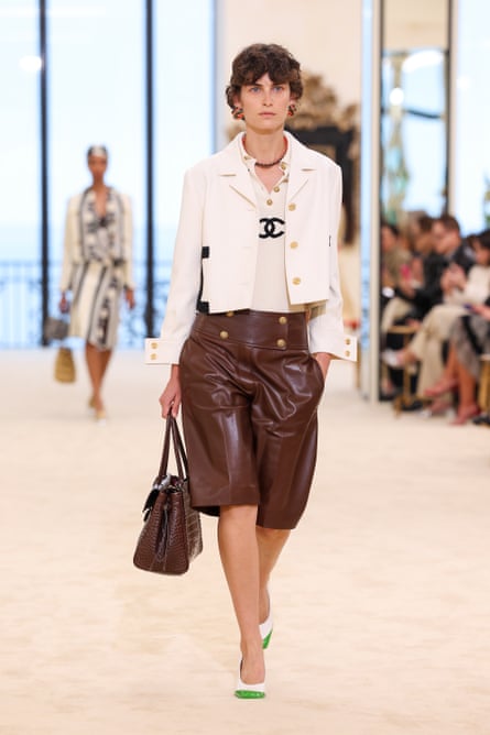 A model wearing a white polo shirt, with two black Cs on the front, a white jacket and brown leather shorts walks the runway