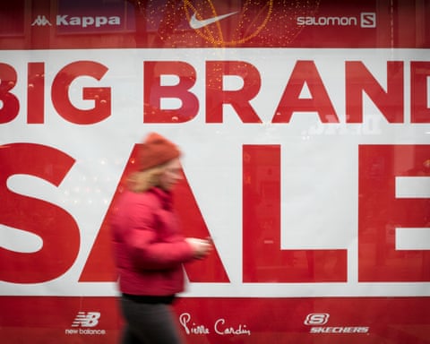a woman in a red puffer jackets passes a shop window with 'big brand sale' in huge red letters