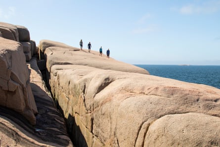 A huge granite cliff by the sea with four walkers in the distance