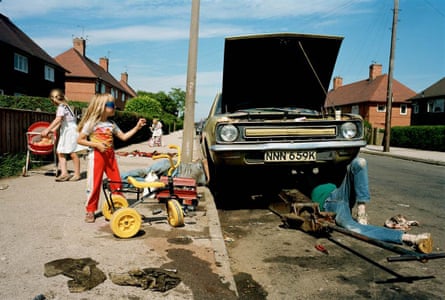 A small girl on a bike stands next to a man working under a car