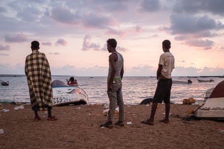 Three men, one wrapped in a checked blanket, look out to sea past small fishing boats.