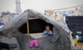 A Palestinian child, about four years old, leans on a wooden frame and looks out of a makeshift tent in a displacement camp: the child has fair hair and wears a blue, pink and purple striped top. This garment, and a pair of bright pink child's trousers hanging to dry on the side of the tent, is in stark contrast to the grey of the tent, which has its roof covered in plastic sheeting, and the grey of the sky in the background. A coil of barbed wire is seen in the foreground.