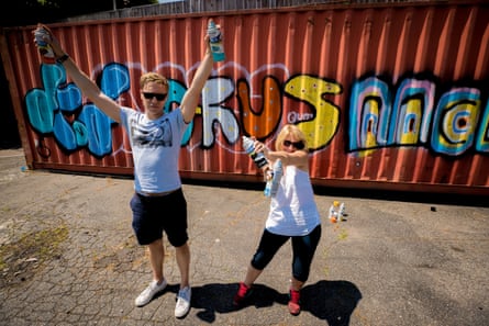 Russell Howard and his mother holding up spray cans in front of colourful graffiti on corrugated iron