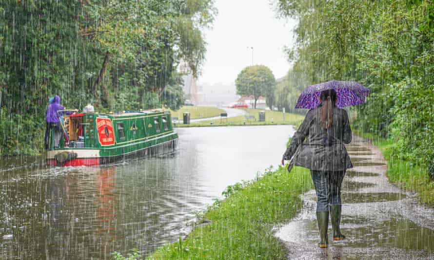 A woman in wellies shelters under umbrella walking on towpath alongside narrowboat in heavy UK rain
