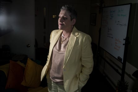 Ashley Dalton stands in her darkened office. There is a whiteboard behind her. She is 53 with short grey hair and wears red lipstick, gold necklaces, a light brown silky top and a pale yellow trouser suit.