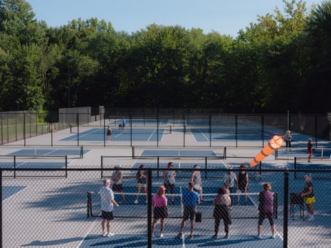 A group of people participating in a pickleball clinic is seen standing in the foreground on pickleball courts while tennis courts are seen in the background. 