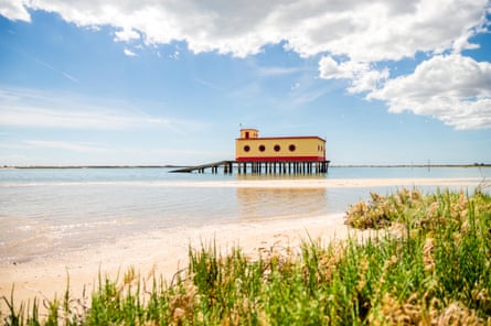 A cream and red lifeguard building on stilts with porthole windows, viewed from a sunny sandy beach fringed with grassy plants