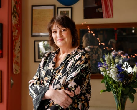 Daniela Nardini, wearing a floral dress, stands, arms folded, in her home, with framed pictures on the wall and a vase of flowers.
