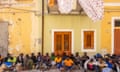 A group of African people sitting on the ground outside a yellow house with wooden shutters in Lampedusa.