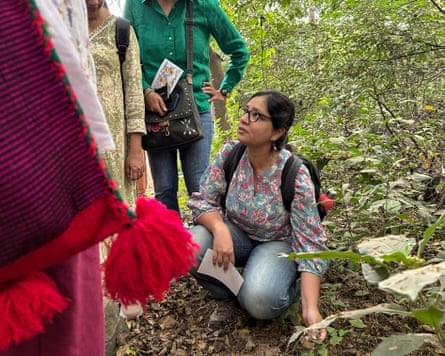 A woman crouches down in a wood and looks up to other women with a plant in her hand