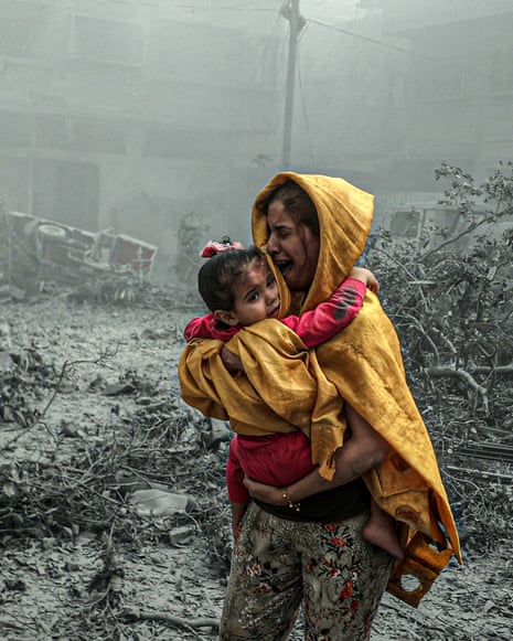 A woman holding a child, both wearing yellow, against a grey background of buildings and debris