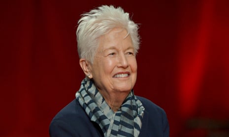 Older white woman, short white hair, scarf, smiling, in front of red curtain.