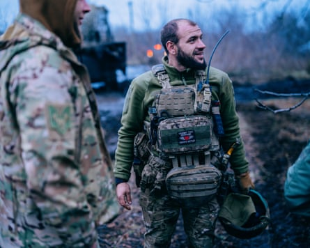 Pavuk, wearing camouflage uniform, is wearing a large camouflage backpack on his front and is holding his helmet in his hand, he is looking to the side, talking to someone off-camera