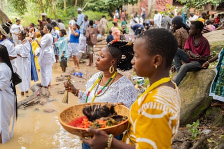 A crowd stands by a river’s edge, with a young African woman holding a wooden bowl in the foreground with what looks like flowers and fruit with an older woman next to her