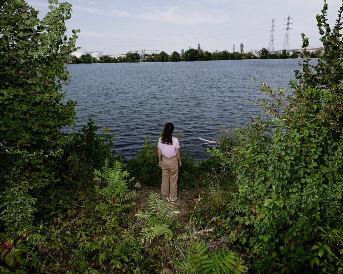 Duguy, with back to camera, stands amid foliage looking across to the other side of the Rhône where a chemical factory is based
