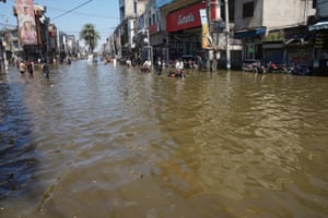 People wade through knee-deep floodwater on an urban street, two of them pushing motorbikes