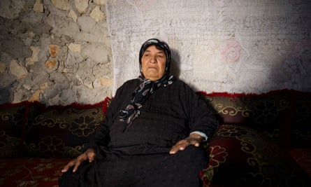 A grim-faced older woman in a black headscarf and black dishdasha robe sits in a crudely built building