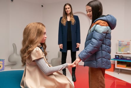 Cady reaches out to touch the doll M3gan, which is sitting on a stool. Gemma watches on, smiling
