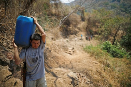 A Latino man carries a heavy plastic jerrycan on his shoulder along a dry hillside