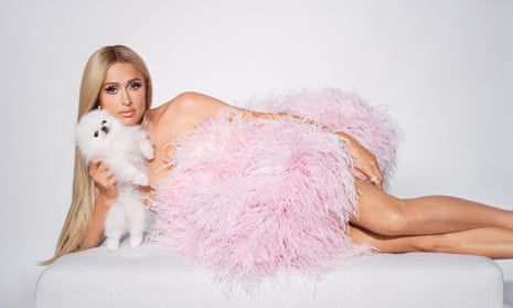 Paris Hilton lying on a couch with a small white fluffy dog and wearing a pink feather dress