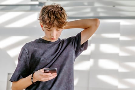 Teenage boy is focused on using his smart phoneThe teenager staring intently at the mobile phone he holding in one hand while holding the back of his head with his other hand while standing against a white wall with shadows on it