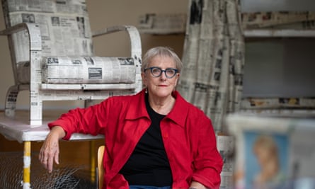 Bobby Baker at Tate Britain, 2023, wearing bright red jacket, black top and blue-framed glasses, sitting in front of a newspaper-covered chair in her installation