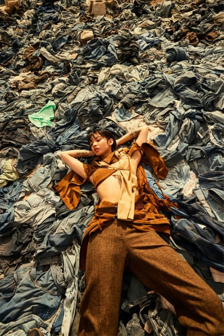A model wearing brown trousers and a crop top lies on a pile of dumped clothes