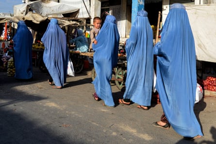 Five Afghan women, dressed in powder blue burqas, walk away from the camera in a line on a street at a market in Kandahar. One woman in the center holds a toddler.
