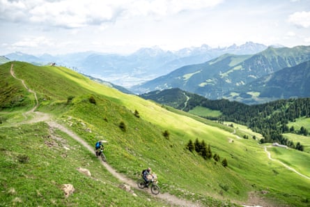 Two cyclists on a trail on a grassy hillside with a valley and mountains in the background