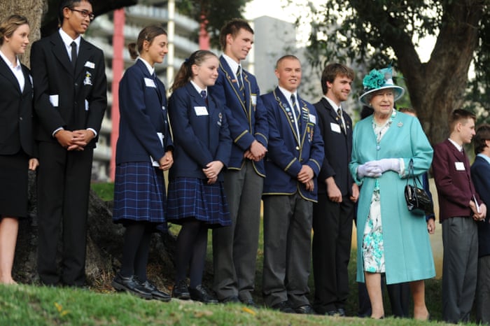 Queen Elizabeth II walks past a line of school students in uniform. She is wearing a teal blue coat and hat