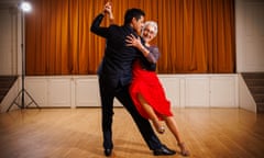 Victoria Zaragoza-Martinez, in glasses, a lace dress and high heels, laughs as she leans against Argentinian instructor Adriàn Pèrez in a tango dance move on a wooden floor in front of a curtained stage