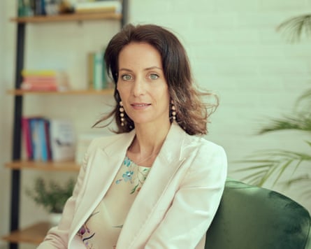 A woman in a white jacket over a pale blouse with small flower motifs sits in a room with a book shelve in the background