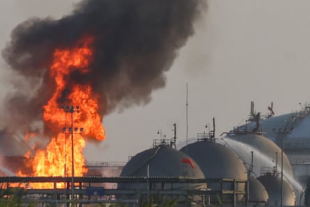 Flames and black smoke rise above an oil facility with tall storage domes to one side.