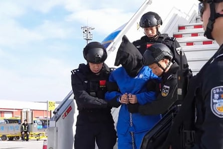A handcuffed man with a bag over his head is led down stairs from an aircraft