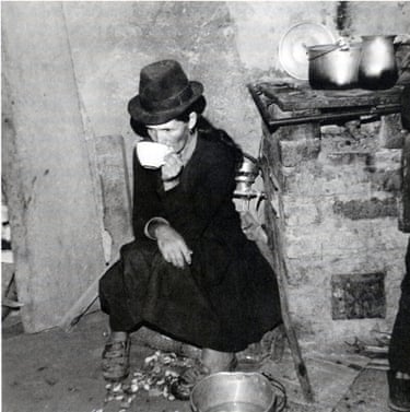 a woman wearing a hat sits drinking coffee by a kitchen stove