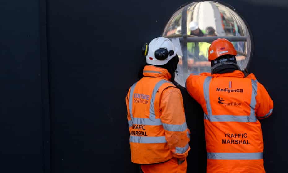workers in orange work overalls with the Carillion logo peer through a construction site hoarding