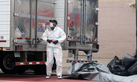 A dressed in a hazmat suit and face mask stands in front of the doors of two big refrigerated trucks outside a funeral home in Brooklyn, New York.