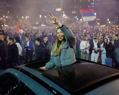 A woman leans from a car’s sunroof making a peace sign amid a celebrating crowd in Belgrade