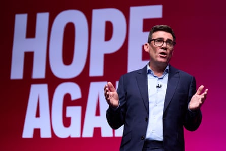 Andy Burnham speaks at the conference. He has his hands raised and outstretched, and stands in front of white lettering reading Hope Again on a pinkish red background. He wears glasses and a dark blue suit with an open-necked pale blue shirt.