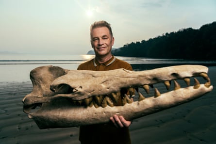 A smiling man holding huge skull of a whale in front of darkening sky and sea