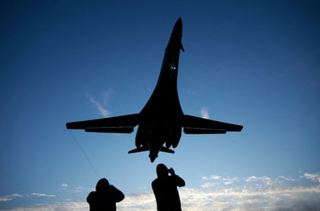 दो लोग, नीले आकाश की छाया में, ऊपर देखते हैं और एक बड़े बमवर्षक हवाई जहाज की तस्वीरें लेते हैं, जो नीले आकाश की छाया में, सिर के ऊपर उड़ रहा है।