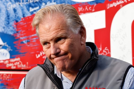 Man in grey vest makes an expression while standing outside in front of red, white and blue background