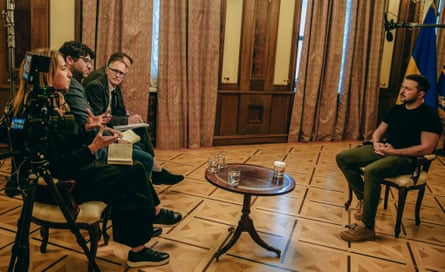 Viner, Walker, Hopkins and Harding sit in a row, facing Zelenskiy. A low table is between them with glasses of water on. A camera next to Viner is filming the interview and there is a microphone over Zelenskiy’s head. Floor-length curtains cover windows in the background. A large Ukrainian flag can be seen in the corner of the room.