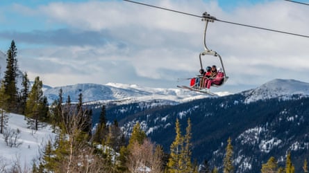 Four skiers sitting on a chairlift above snowy forest with mountains behind